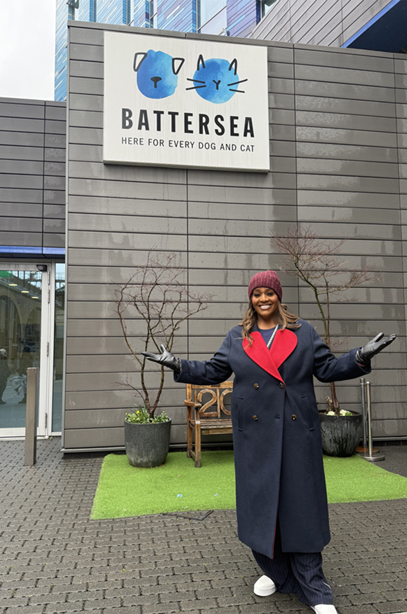Alison Hammon standing with her arms open and smiling under the Battersea dogs and cats home sign