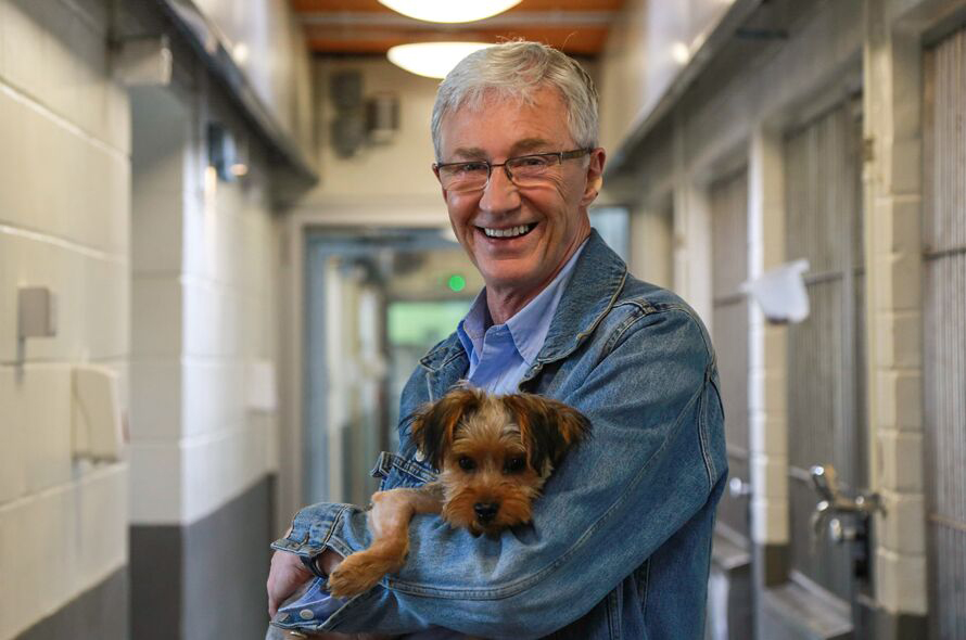 Paul O'Grady stands in the corridor of a Battersea kennel holding a small brown dog