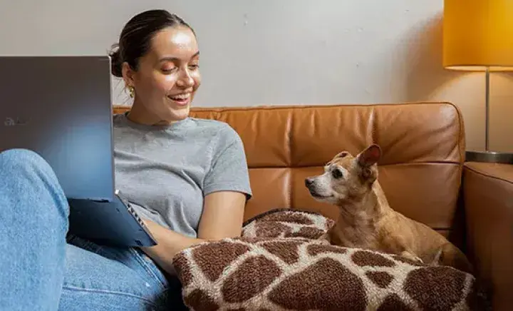 A fosterer sits with a laptop on sofa with a small foster dog next to her 